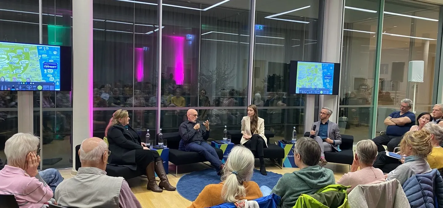 Menschen sitzen im Halbkreis um ein Podium mit vier Personen