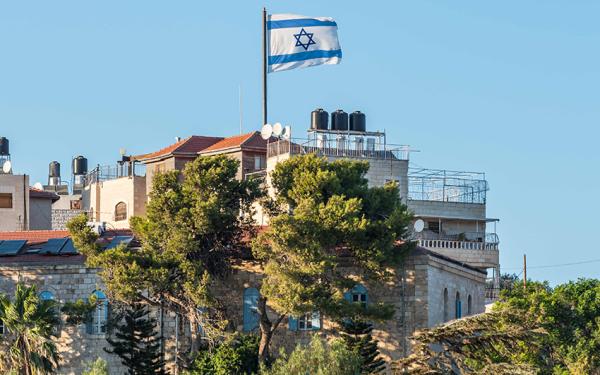 Eine Israel-Flagge weht auf Wohngebäuden, die an einem Steilhang stehen, der mit Bäumen bewachsen ist.