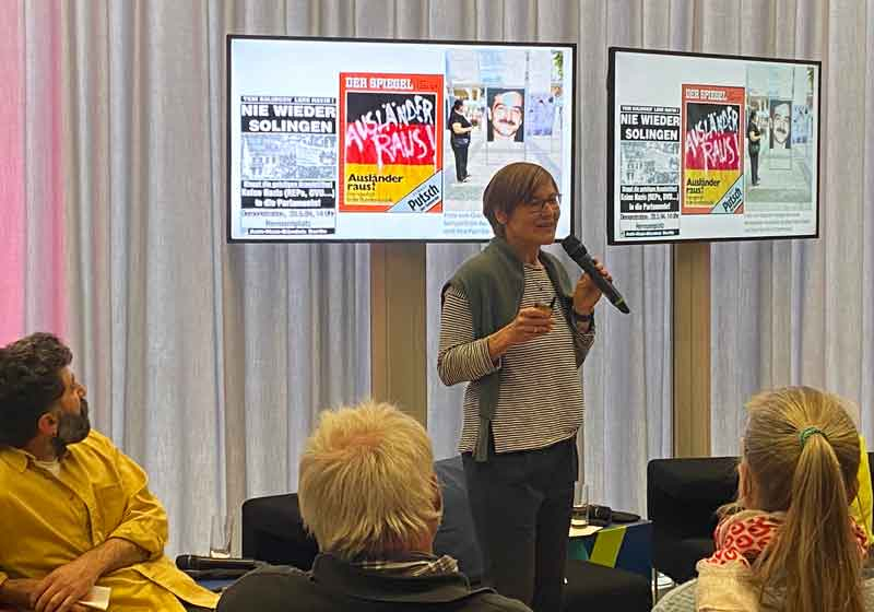 Eine Frau mit Mikrofon steht vor zwei großen Bildschirmen, vor ihr sitzt Publikum