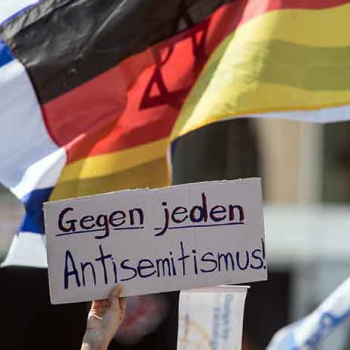 Das Bild zeigt eine Hand, die ein Pappschild hochhält. Darauf steht geschrieben "Gegen jeden Antisemitismus". Im Hintergrund sieht man die deutsche und die israelische Flagge.